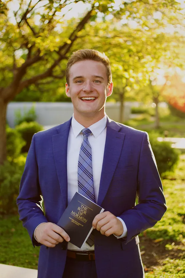 Young man in a navy suit holding scriptures, smiling in afternoon sunlight