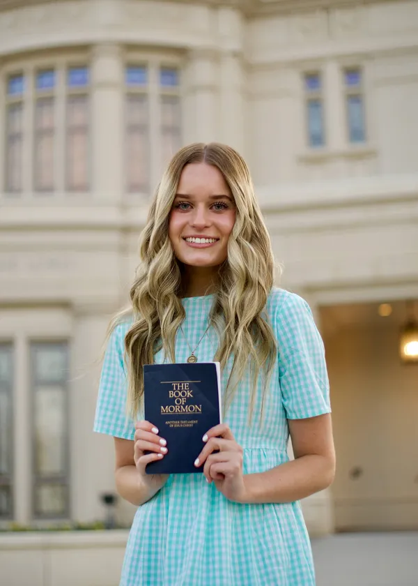 Young woman in a missionary dress holding the Book of Mormon in front of a temple