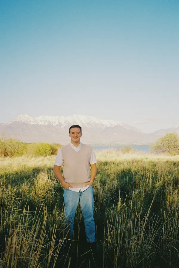 Young man in tall grass with lake and snow-capped mountains behind him, film look